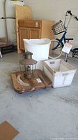 Photo of vintage oil lamp on wooden furniture mover with two white plastic bins and a white waste paper basket visible, as well as wooden cabinet and blue mobility aid.