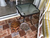 Glass top metal table with bamboo and wrought iron plant stands on tiled patio floor near sliding glass door.