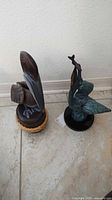 Photo showing two pelican figurines side by side on tile floor against wall. Left figurine is carved wood with rope base, right figurine is heavy metal with Verdi Gris finish on black base.