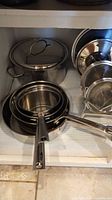 Stacked stainless steel saucepans and frying pans with handles visible, showing shiny metallic surface.