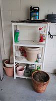 White plastic shelving unit with flower pots, gardening chemicals, and tools arranged on and around it.