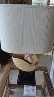 Front view of lamp showing cream-colored textured fabric shade and ceramic shell base on black rectangular stand.
