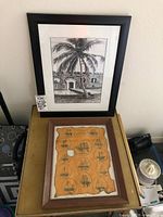 Two framed vintage art pieces on a table: one print of a Key West fort with a palm tree in front, and one antique style nautical poster from the Wreckers Museum.