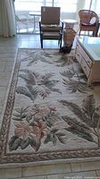 Full view of the ivory area rug featuring large tropical leaves and floral patterns placed in a living room with wicker furniture and tiled floor.