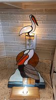 Full view showing both stained glass pelican items, the night light is plugged in and glowing, the larger decor pelican sits on a wooden base with a light bulb illuminating below.