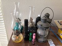 Six vintage oil lamps including four glass and ceramic oil lamps with visible chimneys and metal wick holders, two metal candle lamps with glass globes and handles. Three flashlights of different sizes and colors placed around the lamps. Black bottle of lamp oil behind lamps.