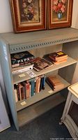 Front view of the solid wood bookcase with books on three shelves and two framed paintings resting on top.