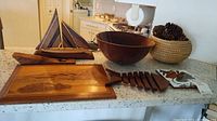 Lot displayed on kitchen counter showing wooden sailboat, wooden bowl, wooden cutting board, fish ceramic trivet, and woven basket with pine cones.