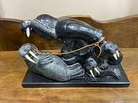 Stone sculpture showing three walruses carved in dark stone with white tusks connected by a leather strap, mounted on a rectangular base.