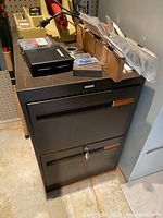 Full view of black metal 2-drawer file cabinet with keys in lock, showing overall condition and size compared to adjacent wall and floor.