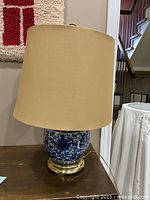 Side view of the table lamp showing blue and white ceramic base, brass base, and beige fabric shade with minor wear visible.