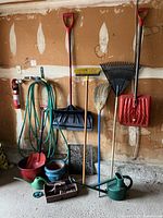 Wide view showing all garden and lawn tools arranged against wall, including shovels, rakes, brooms, hose, watering cans, pots and toolbox with small tools on floor.