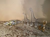 Four glass animal sculptures on a granite surface: two clear giraffes, one colorful spotted fish, and one clear rabbit with a blue collar.
