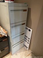 Front angle view of grey metal file cabinet showing four drawers with label holders and recessed handles, cabinet standing beside wall with some adjacent furniture.