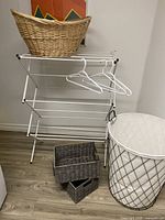 Photo showing the drying rack, natural wicker basket on top, two dark brown wicker baskets stacked on floor, white plastic hangers on drying rack