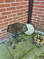 Metal table with black ornate scrollwork base and hexagonal clear glass top, shown with concrete sun ornament and other decorative items on patio corner.