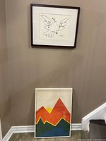 White framed abstract geometric mountain art print in red, orange, yellow, and green colors on white background, leaning against wall near staircase.