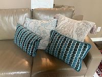 Four decorative pillows arranged on a leather couch: two rectangular teal pillows with geometric patterns, one square beige pillow with teal embroidery, and a large square beige pillow with tassels.