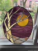 Full view of the stained glass panel hanging in a window, shows the tree and yellow sun with a purple background.