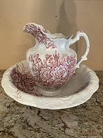 Full view of white ceramic Victorian-style pitcher with red floral pattern sitting inside matching bowl on counter