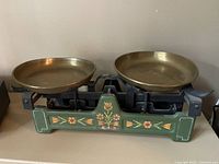 Front view of the vintage DOFT scale showing the green base with floral painting and two brass weighing pans.