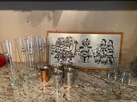 Photo showing six tall Ueli Bier beer glasses with matching decorative logos, four small metal shot cups, one small copper cup, and framed artwork behind them.