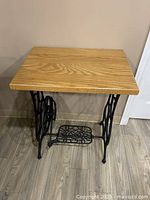 Front angled view of the sewing machine leg table showing the wooden top and decorative cast iron base with foot pedal.