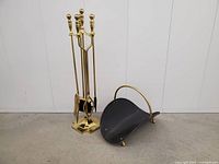 Full view of brass fireplace tools on stand and black metal wood holder with brass handle positioned beside each other on floor.