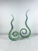 Two green spiral glass sculptures with extended curls and pointed tips, shown side by side on a white surface against a plain background