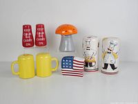 Front view of all 8 assorted ceramic salt and pepper shakers in the lot: red keep calm text, orange mushroom, yellow mugs, chef cartoon, and American flag designs.