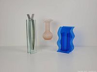 Three vases side by side: clear rectangular MoMA Ribbon Vase with aluminum ribbons inside, pink frosted ribbed glass vase with ruffled top, and blue abstract acrylic vase with wavy edges.