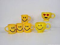 Six vintage yellow smiley face mugs including standard smiley faces, a heart eyes emoji mug, and an oversized Teleflora planter mug.