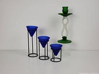 Photo showing the Italian frosted green glass candle holder with twisting stem and three cobalt blue glass candle holders on black metal stands at graduated heights arranged on a clear acrylic riser.