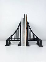 Front view of two black metal suspension bridge design bookends holding several books upright on a flat surface in a minimal setting.