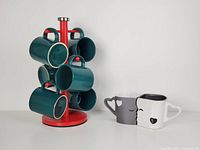 Photo showing the set of 6 dark green ceramic mugs hanging on a red metal tree stand next to the set of 2 white and grey kissing mugs.