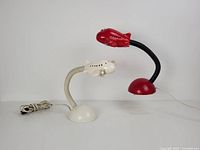 Two ceramic airplane table lamps, one red and one white, shown side by side with cords visible. Both have dome bases and flexible necks supporting airplane-shaped lamp heads.