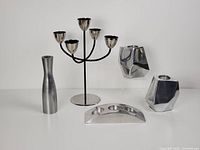 Four chrome metal candle holders on a white background: cylindrical vase-like holder, 5-arm candelabra with round base, small geometric tealight holder, and angular Gotta Design candlestick holder.