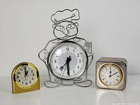 Three vintage clocks on a white surface: chef wireframe clock in center, yellow gold clock left, silver plated silver company clock right