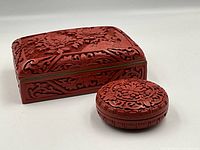 Two cinnabar lacquer lidded boxes, one rectangular with brass and blue enamel accents, and one round with intricate floral carving, shown from an angled front view.