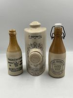 Front view of 2 brown and cream Hamilton stoneware bottles and a white and brown Eaton's stoneware hot water bottle with black lettering.