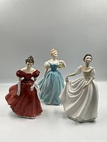 Three Royal Doulton figurines grouped, showing full front views of each in detailed dresses with varying colors.