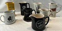 Overview of 8 ceramic advertising pub pitchers featuring multiple Scotch whisky brands in various colors including black, white, yellow. All pitchers have logos and branding related to Scotch whisky.