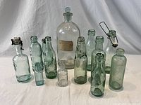 Full lot of 13 antique glass bottles in clear and light green tinted glass with wire and cork stoppers, shown on white background