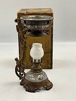 Photo of the Vapo-Cresolene vaporizer with brass ornate base and glass parts, standing in front of the original box.