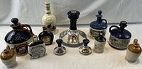Full set of 12 stoneware alcohol decanters shown from front view arranged in two rows on white cloth background.