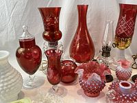 Close-up image of cranberry glass vases with etched floral patterns, hobnail milk glass pieces, small cranberry covered urn, and cut clear glass serving dishes. Also includes miniature kerosene lamps and cranberry hurricanes with brass bases.