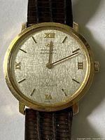 Close front view of linen-textured dial, applied gold indices and slender hands; visible surface scratches on crystal