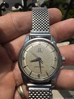 Front view of Omega Seamaster watch held in hand showing patinated cream dial, dauphine hands, applied Arabic numerals and stainless steel mesh band