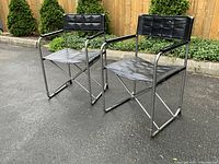 Two chairs at a front right angle on asphalt with wooden fence and greenery in background.