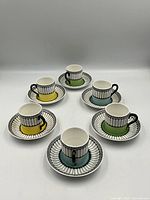Overhead angled photo showing all 6 demitasse cups and saucers arranged in two rows with colored bases in yellow, green, and blue and matching saucers